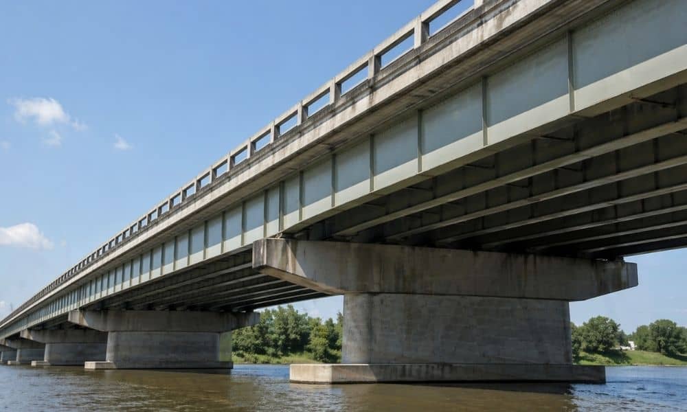 Steel plate girder bridge viewed from below showing steel beams supporting a highway span over a river in daylight.